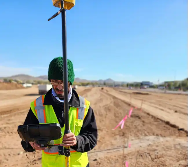A construction worker in a reflective vest holds a surveying instrument on a long, cleared dirt road marked with pink flags under a clear sky